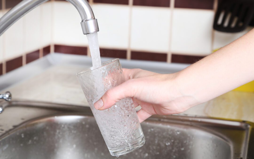 A hand holding a clear glass under a running kitchen faucet, filling it with water, with a tiled backsplash visible in the background.