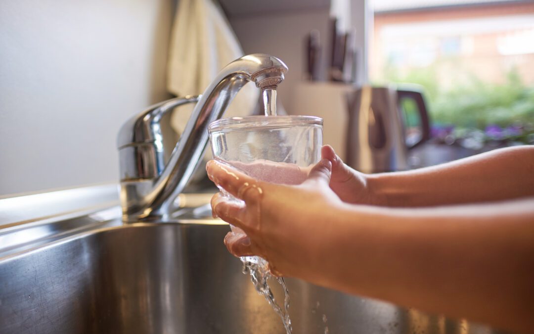 Hands holding a glass under a running faucet in a kitchen sink, with water flowing into the glass. The background features a blurred view of a countertop and a window overlooking greenery.